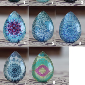 May include: Ten different glass cabochons with various patterns. The cabochons are teardrop shaped and feature designs such as peacocks, mandalas, and floral patterns. The cabochons are clear glass with a variety of colours, including blue, green, purple, and pink.