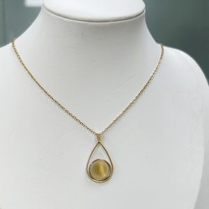 May include: A gold necklace with a fine chain and a teardrop-shaped pendant. The pendant has a circular, textured design within a gold frame. The necklace is displayed on a white mannequin.