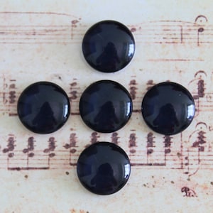 Puede incluir: Cinco botones redondos de vidrio azul marino brillante dispuestos sobre una partitura antigua. Los botones están espaciados uniformemente, con un ligero reflejo visible en sus superficies. El fondo presenta notas musicales descoloridas.