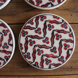K&ouml;nnte beinhalten: Set aus vier wei&szlig;en Keramik-Untersetzern mit einem roten und blauen geometrischen Muster. Die Untersetzer zeigen ein sich wiederholendes Design aus stilisierten Blumen und Bl&auml;ttern.