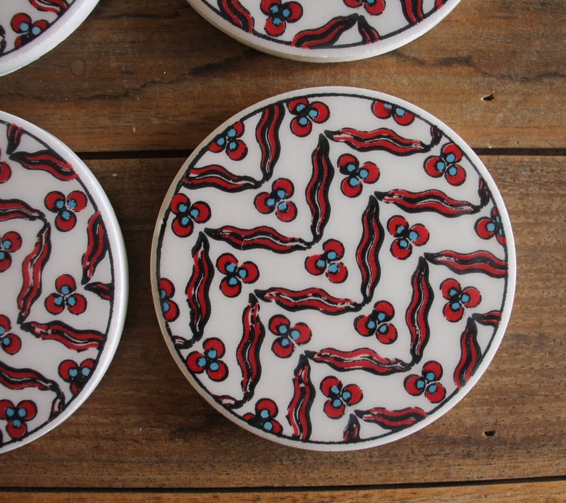 K&ouml;nnte beinhalten: Set aus vier wei&szlig;en Keramik-Untersetzern mit einem roten und blauen geometrischen Muster. Die Untersetzer zeigen ein sich wiederholendes Design aus stilisierten Blumen und Bl&auml;ttern.