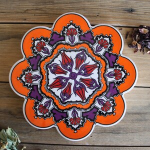 May include: Orange and purple ceramic trivet with a floral design. The trivet is shaped like a flower with eight petals.