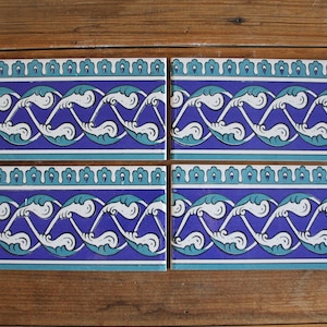 Op de afbeelding: Vier blauwe en witte keramische tegels met een herhalend patroon van gestileerde witte bladeren en spiralen op een blauwe achtergrond. De tegels hebben een turquoise rand.