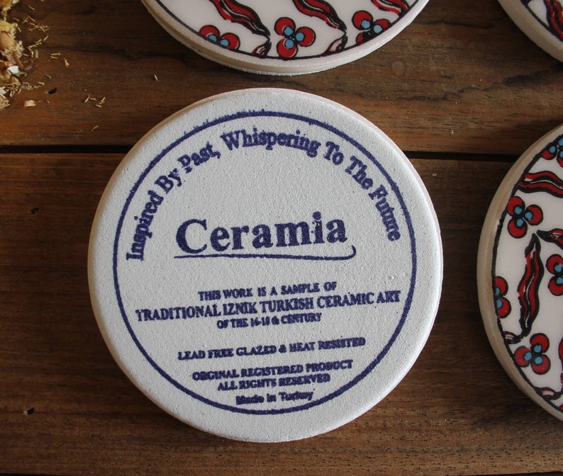 K&ouml;nnte beinhalten: Wei&szlig;er Keramikuntersetzer mit blauem Text, der "Inspired By Past, Whispering To The Future, Ceramia" lautet. Der Text setzt sich fort mit "This work is a sample of Traditional Iznik Turkish Ceramic Art of the 16-18th Century. Lead Free Glazed & Heat Resisted. Original Registered Product. All Rights Reserved. Made in Turkey."