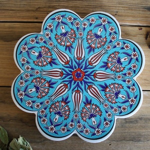 May include: A blue and white ceramic trivet with a floral design. The trivet is shaped like a flower with 10 petals. The design features red, white, and blue flowers and leaves.