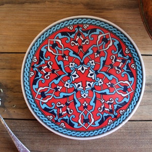 May include: A round ceramic plate with a blue, white and red geometric pattern. The plate has a decorative border with a repeating pattern.