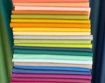 May include: A vertical stack of folded fabric squares in a wide variety of colors including tan, orange, pink, yellow, green, teal, blue, purple, magenta, cream, and black. Each piece of fabric is neatly folded and layered to show the edges.