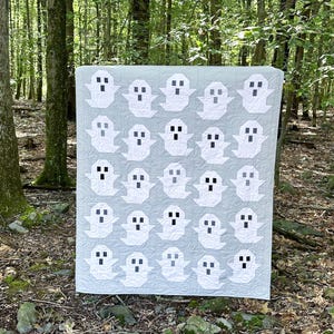 May include: A gray and white quilt with a pattern of ghosts. The quilt is made of fabric squares with a white ghost design on a gray background. The quilt is displayed in a forest setting.