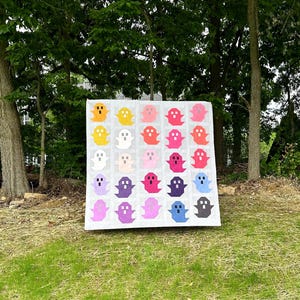 May include: A colorful quilt featuring a grid of ghost shapes in various colors, including yellow, orange, pink, purple, and blue, against a white background. The quilt is displayed outdoors on a grassy area, with trees in the background.