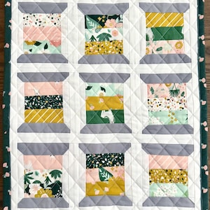 May include: A quilted wall hanging featuring a grid of nine spool-shaped blocks. Each spool is constructed from various patterned fabrics in shades of green, yellow, pink, and white, separated by white sashing and framed by a dark green floral border.