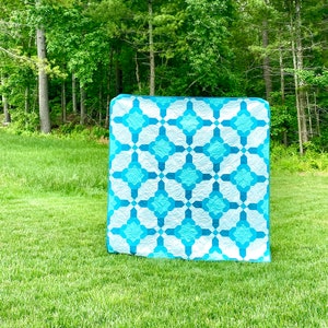 May include: A blue and white quilt with a geometric pattern. The quilt is made of squares and diamonds, and it is folded in half. The quilt is lying on a grassy lawn in front of a forest.