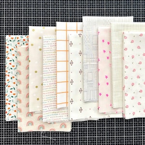 May include: A collection of folded fabric pieces with various patterns. Designs include floral, rainbows, dots, grid, diamond shapes, architectural sketches, stars, and small flowers. Colors are primarily white, cream, pink, orange, and brown.