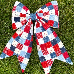 May include: A large, handmade bow featuring a patchwork design in red, white, and blue. The bow is made of fabric squares with various patterns, including polka dots and stars. The bow is set against a backdrop of green grass.