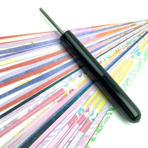 May include: A black paper quilling tool with a thin metal needle. The tool is lying on a surface of colorful paper strips.