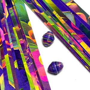 May include: A stack of colorful paper strips with a purple, green, and yellow abstract pattern. Two small, rolled paper beads are in the foreground, one on each side of the stack.
