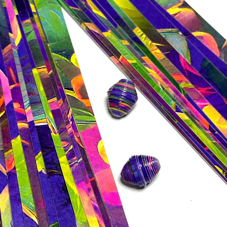 May include: A stack of colorful paper strips with a purple, green, and yellow abstract pattern. Two small, rolled paper beads are in the foreground, one on each side of the stack.