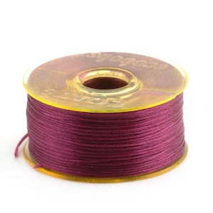 May include: A spool of deep burgundy thread wound around a yellow plastic bobbin. The bobbin has the word "Color" printed on it.