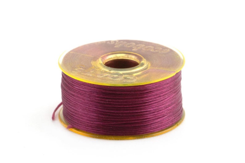 May include: A spool of deep burgundy thread wound around a yellow plastic bobbin. The bobbin has the word "Color" printed on it.
