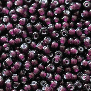 Può includere: Primo piano di una grande quantità di piccole perline di vetro rotonde di colore viola scuro con centri bianchi. Le perline sono disposte in uno schema casuale, creando una superficie testurizzata.