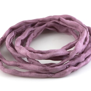 May include: A circular arrangement of twisted, lavender-coloured fabric, possibly silk. The fabric has a soft appearance and a slightly textured surface, suggesting a handmade quality. The colour is a muted purple.