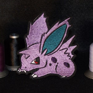 May include: Purple and teal embroidered patch of a cartoon purple creature with a red eye and a spiky back.