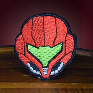 May include: A red and black embroidered patch of a video game character's helmet. The helmet has a green visor and a detailed design.