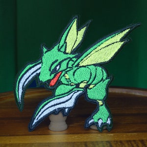 Peut inclure: Patch brodé vert thermocollant représentant une créature de dessin animé avec de grandes ailes et un long museau. La créature a des accents jaunes sur ses ailes et des accents rouges sur son museau.