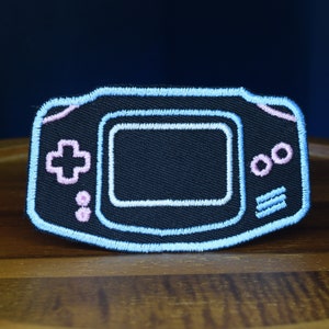 Op de afbeelding: Een geborduurde patch in blauw en roze van een draagbare videogameconsole. De console heeft een zwart scherm en roze knoppen.