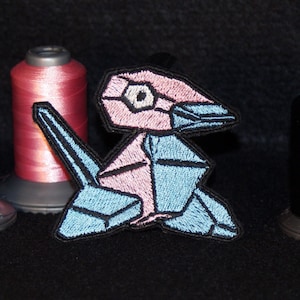 Może przedstawiać: Naszywka w stylu origami w kolorze różowym i niebieskim przedstawiająca stworzenie przypominające ptaka. Naszywka leży na czarnej powierzchni obok trzech szpulek nici w kolorze niebieskim, różowym i czarnym.