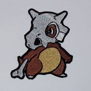 May include: An embroidered iron-on patch featuring a cartoon of a grey and brown Pokemon character with a bone-like tail.