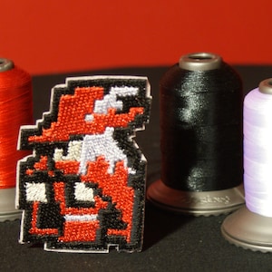 May include: A red, white and black embroidered patch of a pixelated character wearing a red hat. The patch is sitting on a black surface next to three spools of thread: red, black and purple.