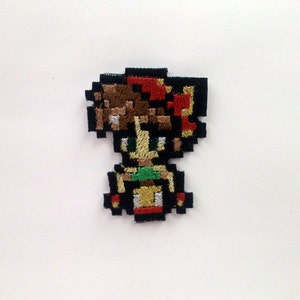 May include: Embroidered patch featuring a pixelated character with brown hair, a red bow, a green shirt, and a yellow skirt. The character is standing with their arms at their sides.