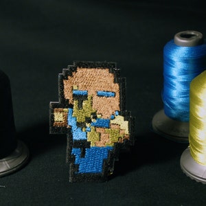 May include: A pixelated embroidered patch depicting a person with brown hair, blue eyes, and a blue shirt. The patch is surrounded by three spools of thread: black, blue, and yellow.