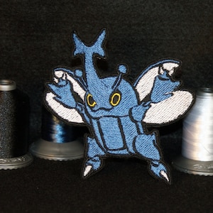 Op de afbeelding: Blauwe geborduurde strijk-op-patch van een cartoon-kever met witte vleugels en gele ogen.