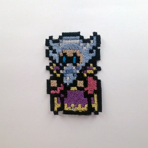 May include: Embroidered patch of a pixelated character with white hair and a blue beard, wearing a purple and pink robe with gold trim. The character is wearing a black hat with a gold trim.