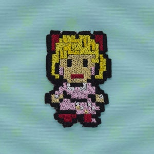 Pode incluir: Etiqueta bordada de uma menina pixelada com cabelo loiro, vestindo um vestido rosa e sapatos vermelhos.