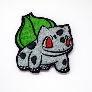 May include: Embroidered iron-on patch featuring a cartoon gray and green Pokemon character with red eyes.