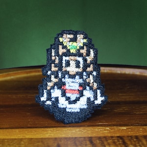 Puede incluir: Parche bordado con un personaje pixelado con cabello negro, una corona verde y una camisa a rayas rojas y blancas. El personaje lleva un vestido blanco y está de pie sobre un fondo negro.