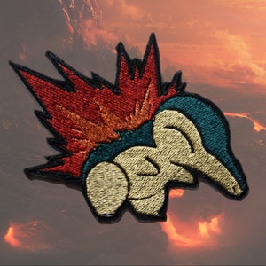 Puede incluir: Un parche bordado con un personaje de dibujos animados con un cuerpo azul y verde y una gran melena roja, naranja y amarilla de punta. El personaje está enroscado en una bola.