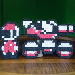 Könnte beinhalten: Zwei gestickte Patches mit pixelartigen Figuren aus einem Videospiel. Der größere Patch ist schwarz, weiß und rot und zeigt eine roboterartige Figur, die eine Waffe hält. Der kleinere Patch ist schwarz, rot und weiß und zeigt eine kleinere Figur mit einem Helm.