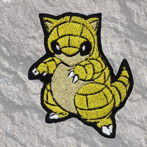 May include: Yellow and brown embroidered iron-on patch of a cartoon creature with a yellow shell and black eyes.