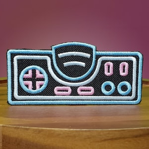 Op de afbeelding: Een geborduurde patch met een retro-videogamesysteem in blauw en zwart met roze knoppen.