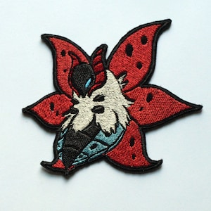 Puede incluir: Parche bordado con una criatura blanca con ojos negros y un diseño de flor roja, negra y azul.