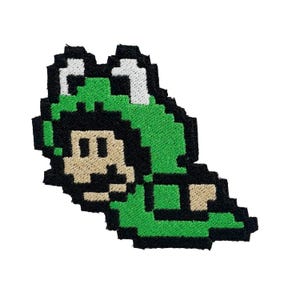 Könnte beinhalten: Gestickter Aufnäher mit einem pixeligen Charakter in einem grünen Froschkostüm. Die Figur hat ein beiges Gesicht, schwarze Augen und eine weiß-grüne Froschkapuze. Der Aufnäher ist schwarz umrandet und vor weißem Hintergrund.