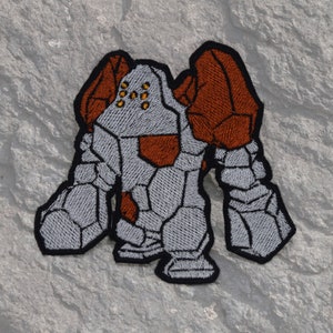 Puede incluir: Parche bordado de un golem de piedra gris y marrón con una superficie texturizada. El golem tiene una cabeza grande con un casco rojo y gris y cuatro pequeños ojos amarillos.