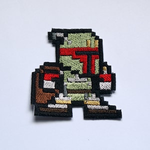 Puede incluir: Un parche bordado de color verde, rojo y marrón que representa a un personaje de Star Wars pixelado, Boba Fett.