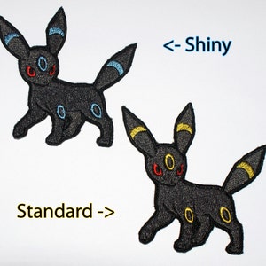 May include: Two embroidered iron-on patches of the Pokemon Umbreon, one with blue accents and the other with yellow accents. The text "Standard ->" is below the blue accented patch and "<- Shiny" is above the yellow accented patch.