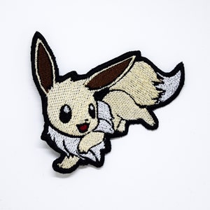 以下が含まれることがあります： ベージュとブラウンの刺繍でできた、ポケモンのキャラクター、イーブイの漫画のキャラクターのパッチ。白いふわふわの尻尾があります。