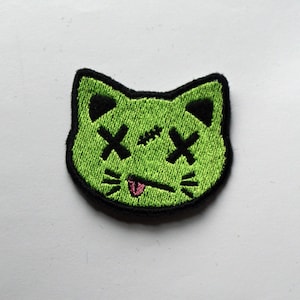May include: Green embroidered iron-on patch of a cat with X's for eyes and a pink tongue sticking out.