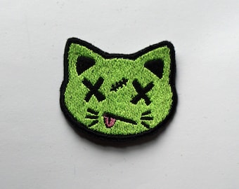 May include: Green embroidered iron-on patch of a cat with X's for eyes and a pink tongue sticking out.